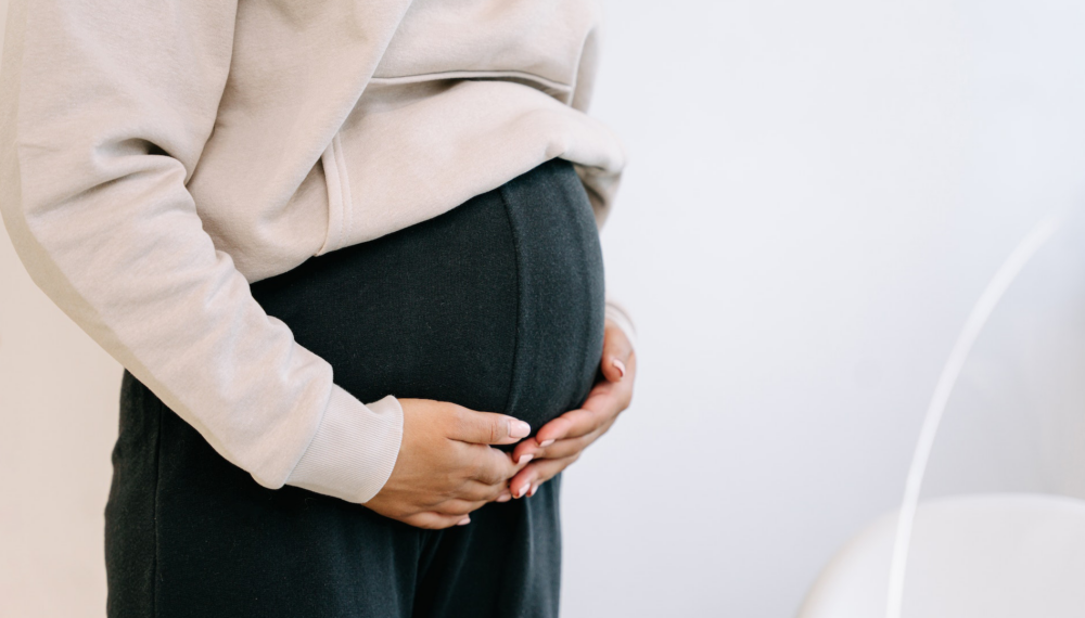 Pregnant woman in black trousers and cream top holding her hands on her belly.