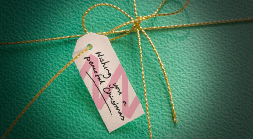 A pink gift tag tied onto a pale green parcel with gold string. The label on the tag features the Tinnitus UK soundwave and text that says 'Wishing you a peaceful Christmas'