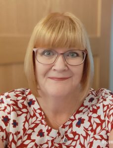 Portrait image of Annette. A woman in her 60's with blonde hair styled in a bob wearing glasses. She has blue eyes and is weating a red top with a white flower pattern on it.