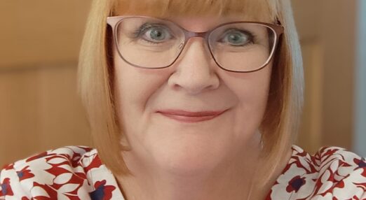 Portrait image of Annette. A woman in her 60's with blonde hair styled in a bob wearing glasses. She has blue eyes and is weating a red top with a white flower pattern on it.