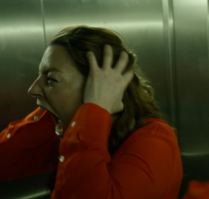 A woman wearing a red shirt screams in a lift holding her ears.