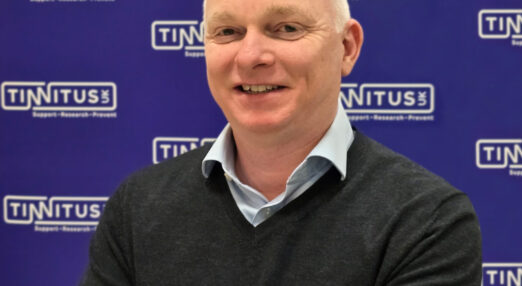 Portrait of CEO Alex Brooks-Johnson in front of a dark blue Tinnitus UK branded background.