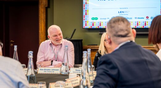 Image shows Alex Brooks-Johnson sitting at a table with other people. There is a screen with CEO's Roundtable behind him.